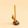 A lit yellow candle is held by the Snake Cake Topper, a handcrafted ceramic holder shaped like a snake wearing a small red and yellow party hat. The charming keepsake is coiled on a pale beige surface that matches the background.