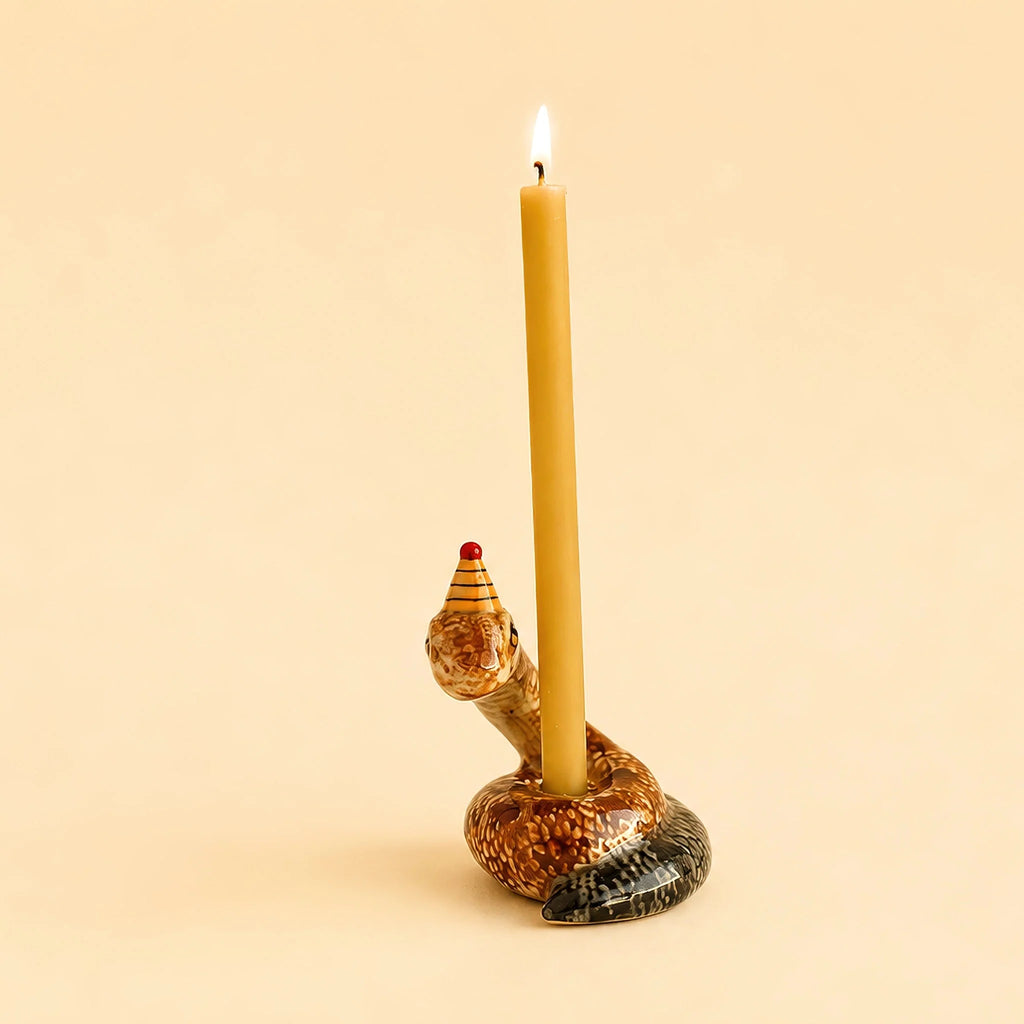 A lit yellow candle is held by the Snake Cake Topper, a handcrafted ceramic holder shaped like a snake wearing a small red and yellow party hat. The charming keepsake is coiled on a pale beige surface that matches the background.