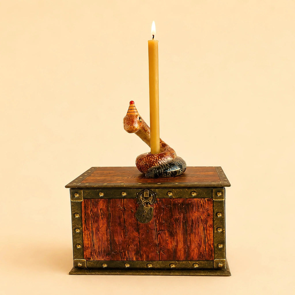 A yellow beeswax birthday candle stands upright, held by the Snake Cake Topper, which sits atop a wooden treasure chest with metal accents and a lock.