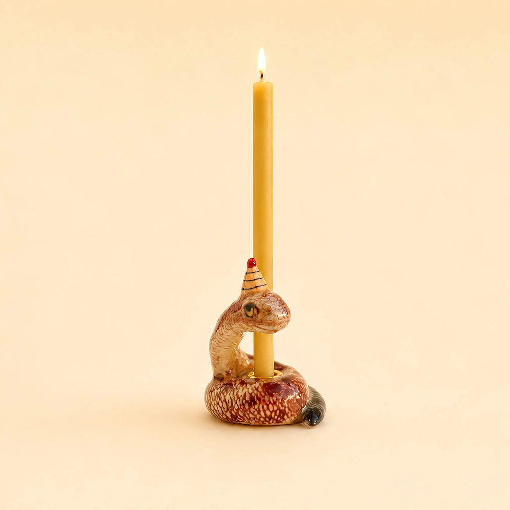 A yellow beeswax birthday candle stands upright in the hand-painted “Snake Cake Topper – Final Sale,” a ceramic candle holder shaped like a coiled snake wearing a small party hat, set against a plain beige background. This whimsical piece makes for a charming collectible.