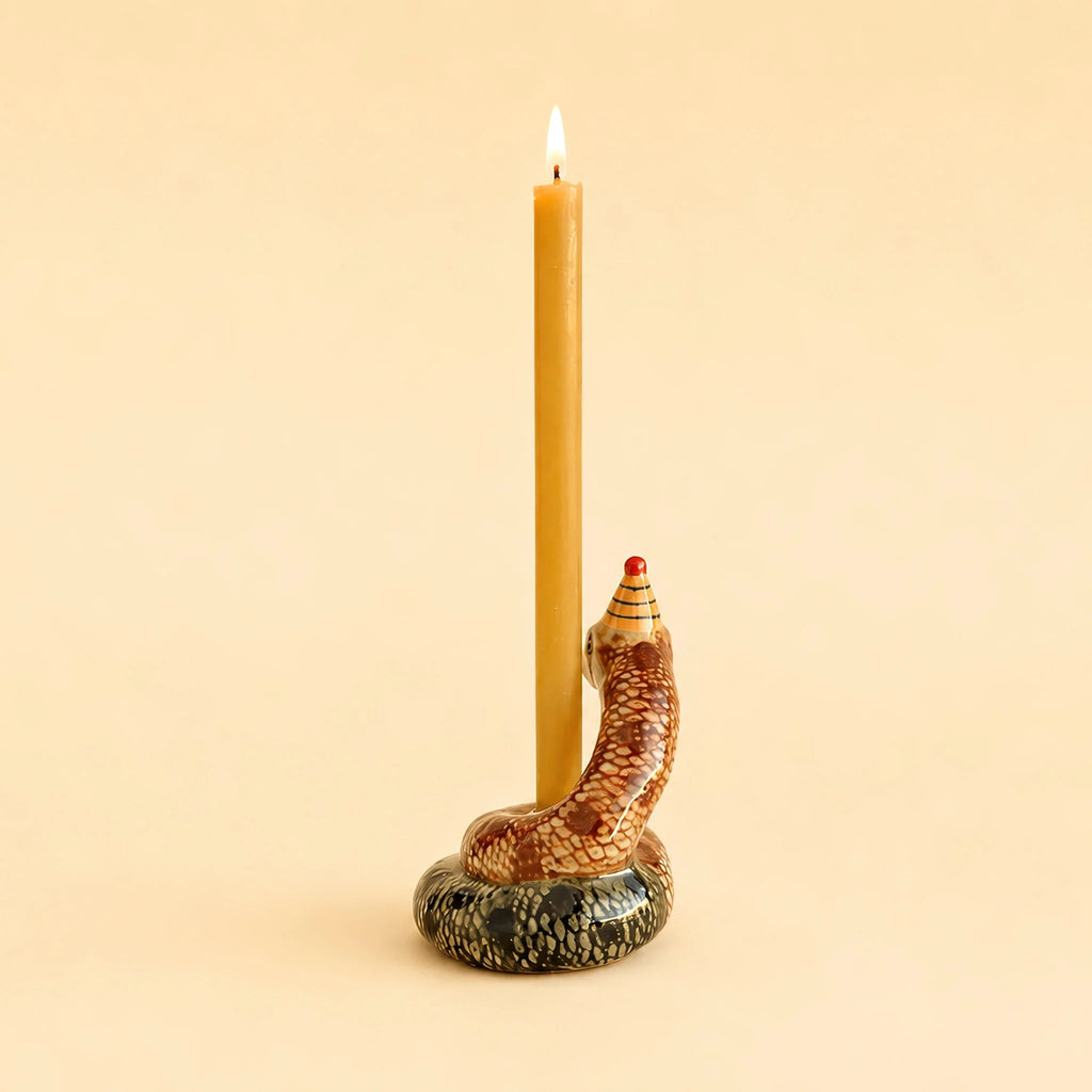 A brown, hand-painted collectible Snake Cake Topper with a red-tipped hood holds a lit, tall, yellow candle against a plain beige background.