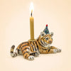 The Tiger Cake Topper - Final Sale is a handcrafted collectible ceramic figurine featuring a tiger wearing a party hat and holding a lit candle on its back, set against a plain beige background.