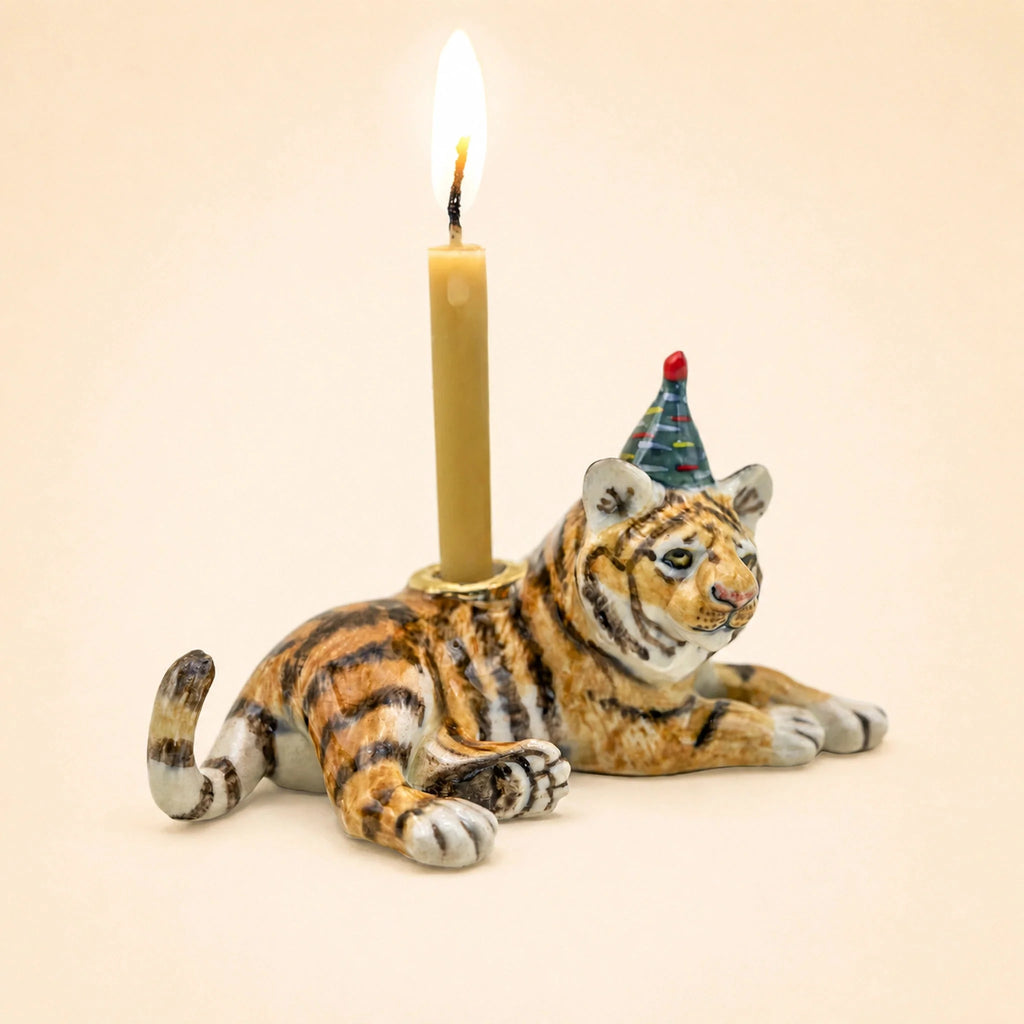 The Tiger Cake Topper - Final Sale is a handcrafted collectible ceramic figurine featuring a tiger wearing a party hat and holding a lit candle on its back, set against a plain beige background.
