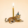 The Tiger Cake Topper - Final Sale is a handcrafted ceramic figurine featuring a tiger adorned with a vibrant party hat and carrying a lit candle on its back, set against a plain beige backdrop—an exquisite collectible ideal for use as an original porcelain cake topper or treasured keepsake.