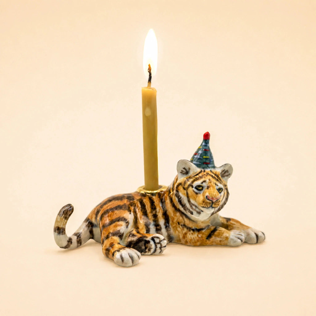 The Tiger Cake Topper - Final Sale is a handcrafted ceramic figurine featuring a tiger adorned with a vibrant party hat and carrying a lit candle on its back, set against a plain beige backdrop—an exquisite collectible ideal for use as an original porcelain cake topper or treasured keepsake.
