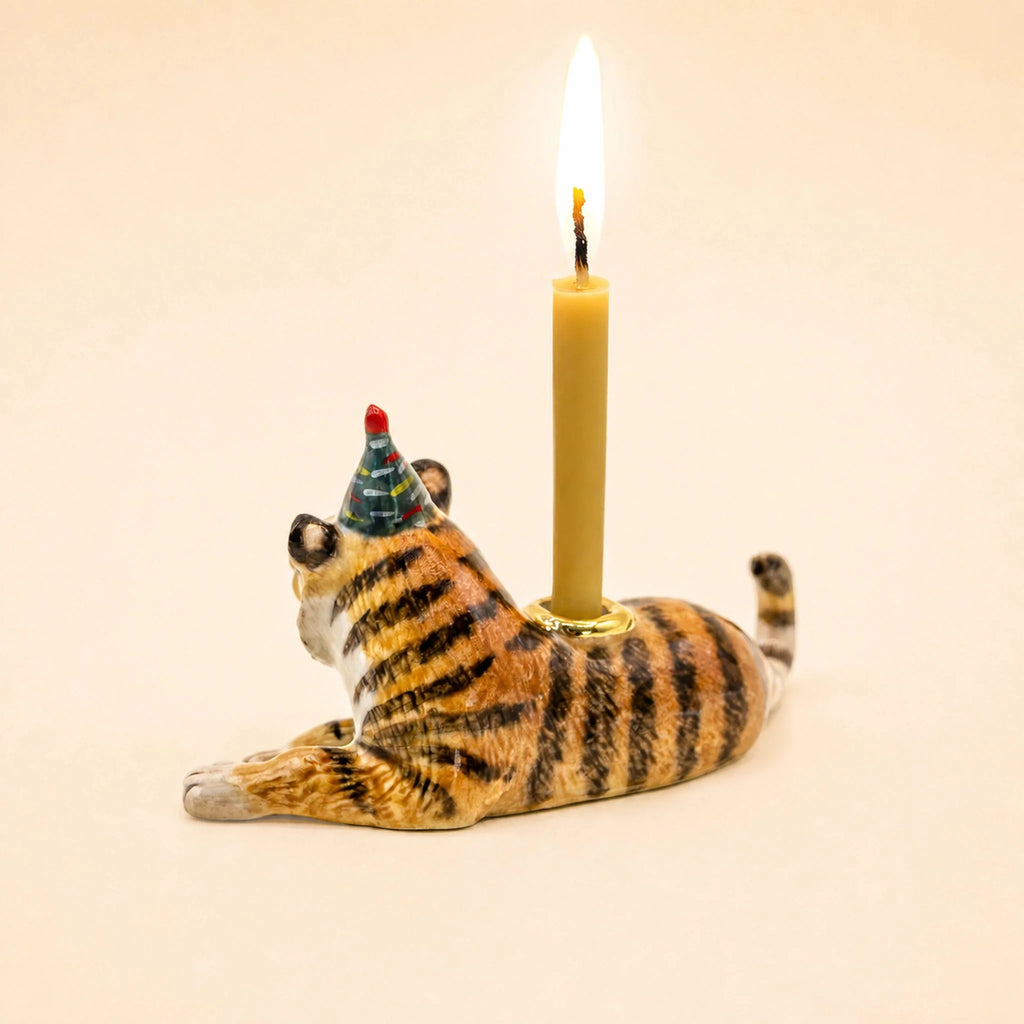 Introducing the Tiger Cake Topper - Final Sale: a handcrafted ceramic collectible featuring a tiger figurine wearing a blue party hat, lying on its stomach and balancing an upright lit yellow candle on its back, displayed against a simple light background.