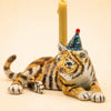 Meet the Tiger Cake Topper - Final Sale: This handcrafted ceramic collectible features a tiger figurine lying on its side, adorned with a blue party hat decorated in red dots. Behind the tiger stands an upright yellow candle, making it a delightful accent or whimsical porcelain cake topper. Set against a soft beige background, this unique piece radiates playful charm and celebrates any occasion with style.