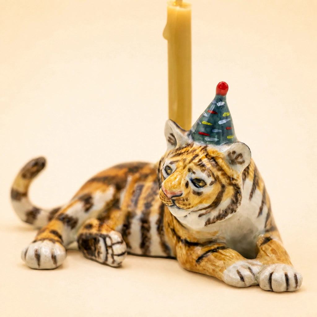 Meet the Tiger Cake Topper - Final Sale: This handcrafted ceramic collectible features a tiger figurine lying on its side, adorned with a blue party hat decorated in red dots. Behind the tiger stands an upright yellow candle, making it a delightful accent or whimsical porcelain cake topper. Set against a soft beige background, this unique piece radiates playful charm and celebrates any occasion with style.