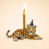 Introducing the Tiger Cake Topper – Final Sale: This handcrafted collectible features a ceramic tiger figurine donning a vibrant party hat and carrying a lit candle on its back, set against a plain beige background. Ideal as a porcelain cake topper or an enchanting addition to your keepsake collection, it brings whimsical flair to any celebration.