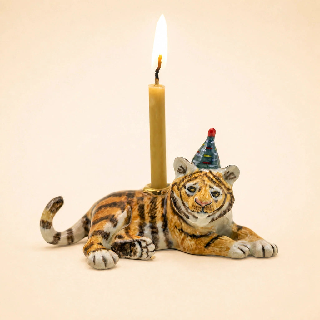 Introducing the Tiger Cake Topper – Final Sale: This handcrafted collectible features a ceramic tiger figurine donning a vibrant party hat and carrying a lit candle on its back, set against a plain beige background. Ideal as a porcelain cake topper or an enchanting addition to your keepsake collection, it brings whimsical flair to any celebration.