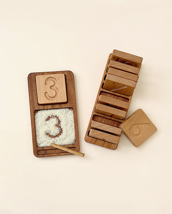 The Wooden Counting & Tracing Tray - Made in USA is an educational tool. One tray shows the number 3 with rice and a wooden pen for tracing, while other trays are stacked nearby, the top displaying the number 6.