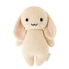 A Cuddle + Kind Baby Bunny with a neutral expression, featuring long pink-lined ears and a pastel cream body, hand-knit from Peruvian cotton yarn. It sits upright against a white background.