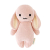 A Cuddle + Kind Baby Bunny hand-knit plush rabbit with long ears, sitting upright. It has stitched eyes and mouth, beige inner ears, and a small fabric tag on one side. The background is white.