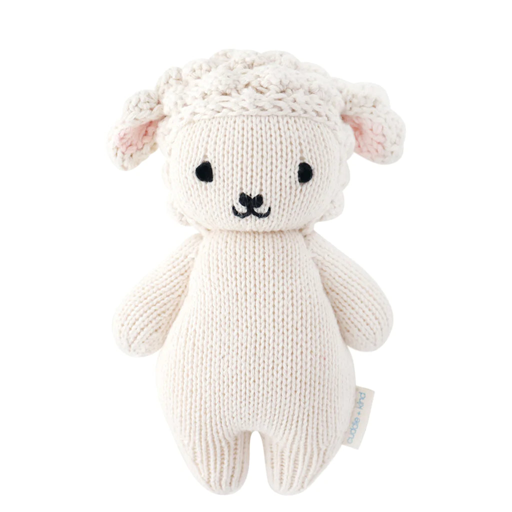 A Cuddle + Kind Baby Lamb, hand-knit from Peruvian cotton yarn, with a white body and cream-colored, curly textured wool on its head, featuring simple black stitched eyes and nose.