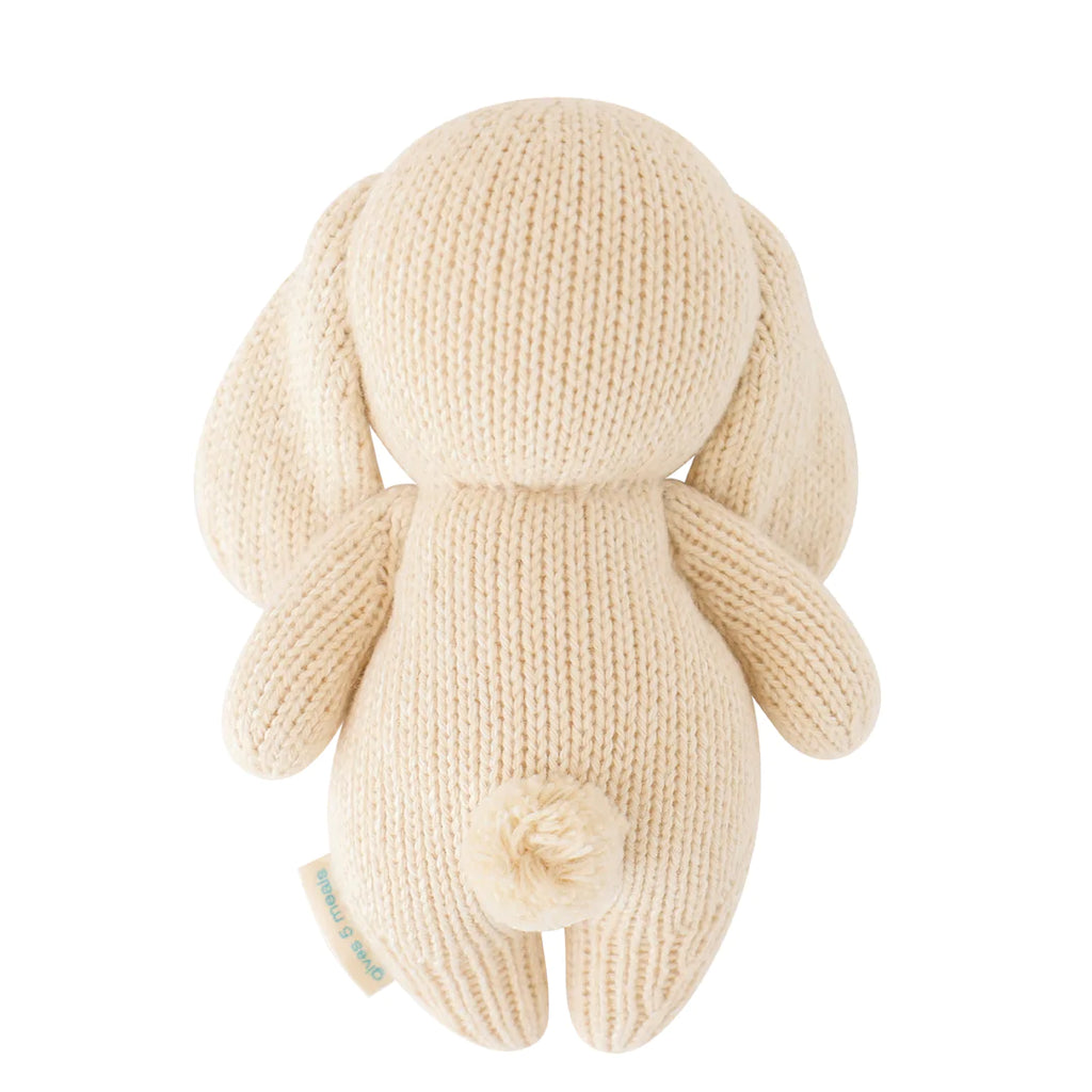 A hand-knit beige Cuddle + Kind Baby Bunny stuffed animal, made of Peruvian cotton yarn, featuring long ears and a rounded tail, shown from the back, against a white background.