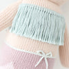 Close-up of a Cuddle + Kind Luna The Mermaid showing intricate details like a light blue fringed band on a pink base, accented with a small white bow. The textures emphasize the craftsmanship of the knitting.