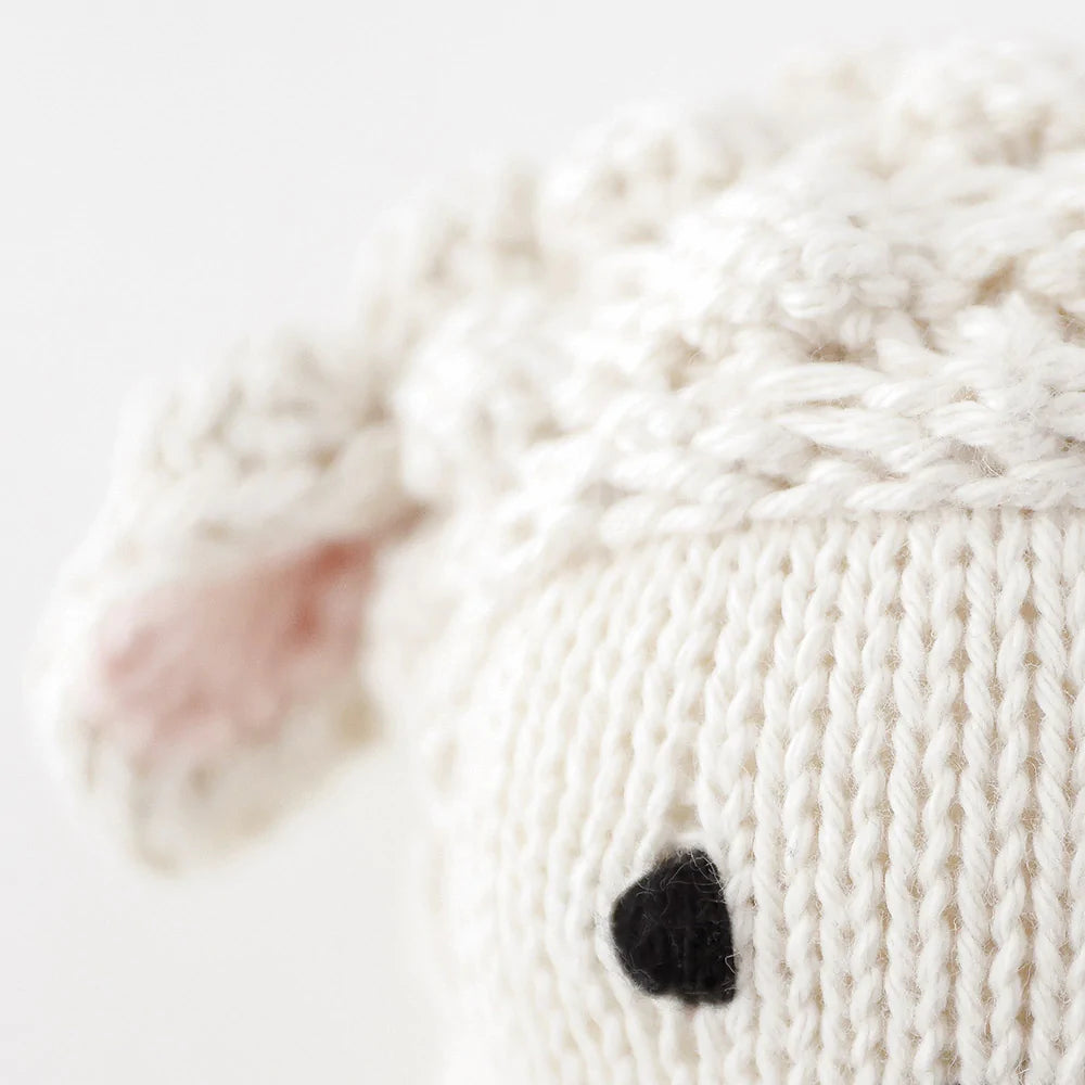 Close-up of a hand-knit Cuddle + Kind Baby Lamb made from Peruvian cotton yarn, with visible stitched details, featuring a soft white texture and distinct black and pink accents for the eyes and ears.