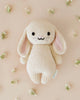 A Cuddle + Kind Baby Bunny, hand-knit from Peruvian cotton yarn, with long ears and a smiling face, lying on a light pink background surrounded by small white flowers.