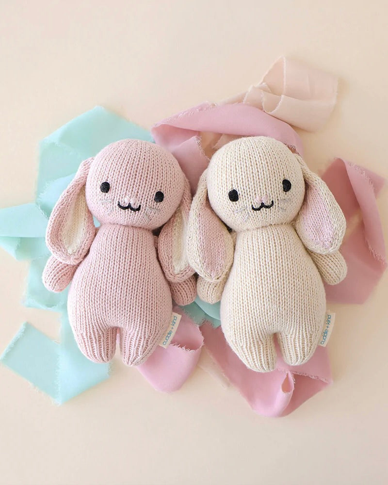 Two adorable Cuddle + Kind Baby Bunnies, one pink and one cream, are lying on a soft pastel background of blue and pink fabric scraps.