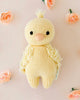 A hand-knit Cuddle + Kind Baby Duckling plushie with soft, textured wings and a tuft of hair on top, surrounded by pink roses on a light pink background.