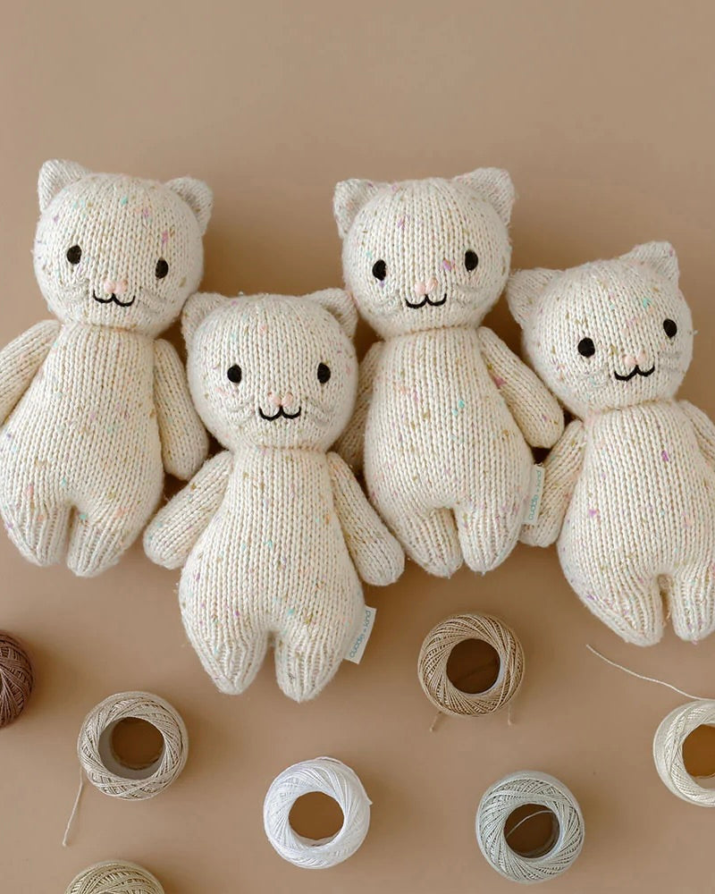 Four Cuddle + Kind Baby Kittens displayed in a row against a beige background, with visible yarn spools in front. Each bear has a smiling face and specks of color in their fabric.