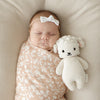 A sleeping newborn baby with a white bow headband, snugly swaddled in a floral pink blanket, lying next to a Cuddle + Kind Baby Lamb toy on a beige textured surface.