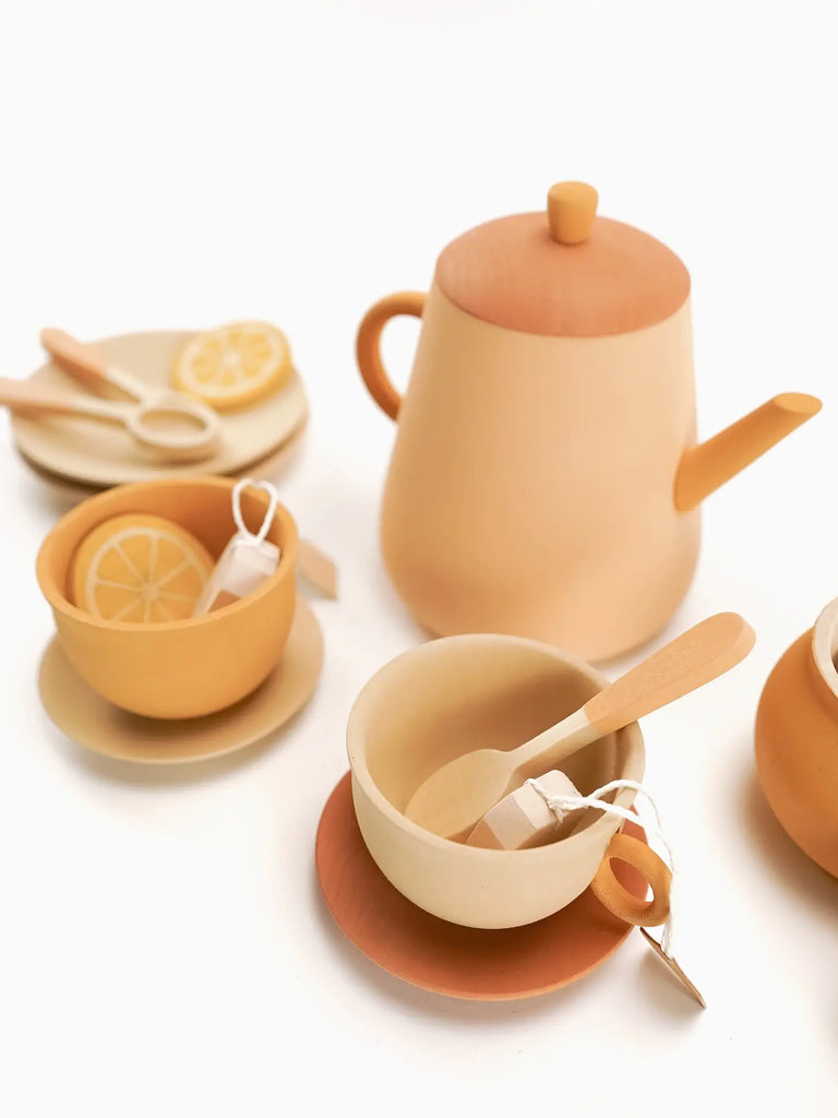 A collection of ceramic kitchenware in a soft peach color, including a Handmade Wooden Tea Set - Flower, cups with lemon slices, small bowls, and wooden spoons, arranged on a light background.