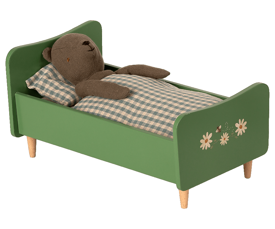 A Teddy Dad - Dusty Green tucked into a child's wooden bed with green frames, handpainted details, and floral decoration. The bear is lying on a checkered blanket.