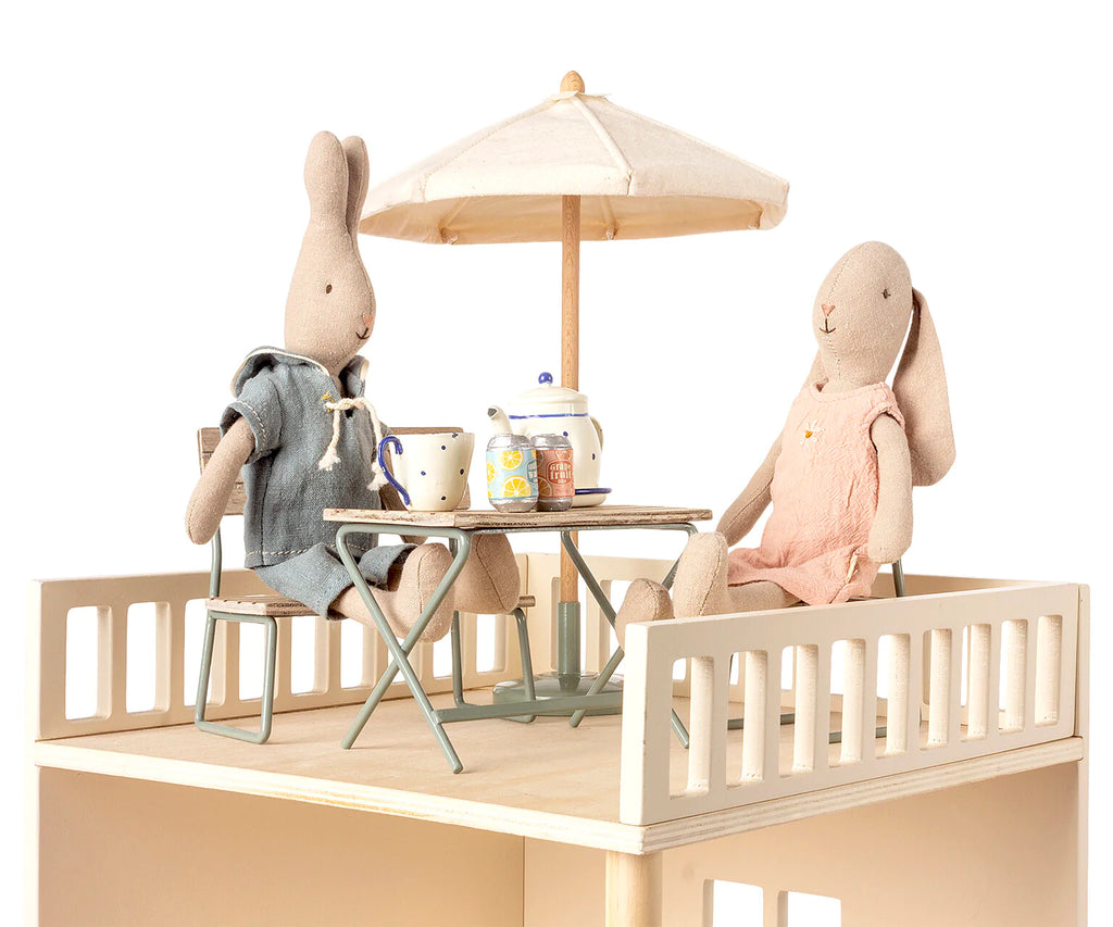 Two fabric bunny dolls sit at a small patio table under a Maileg Miniature Sunshade, enjoying a tea party. One bunny wears a blue-gray outfit, while the other wears pink. The setting is on a cozy miniature garden set with a white railing, perched on an enchanting rooftop terrace.