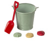 A green bucket and a red shovel from the Maileg Sand Toys set are placed next to three toy seashells in yellow, green, and red. The bucket has a handle and the word "Maileg" embossed on it—a perfect sand toy for your vacation.