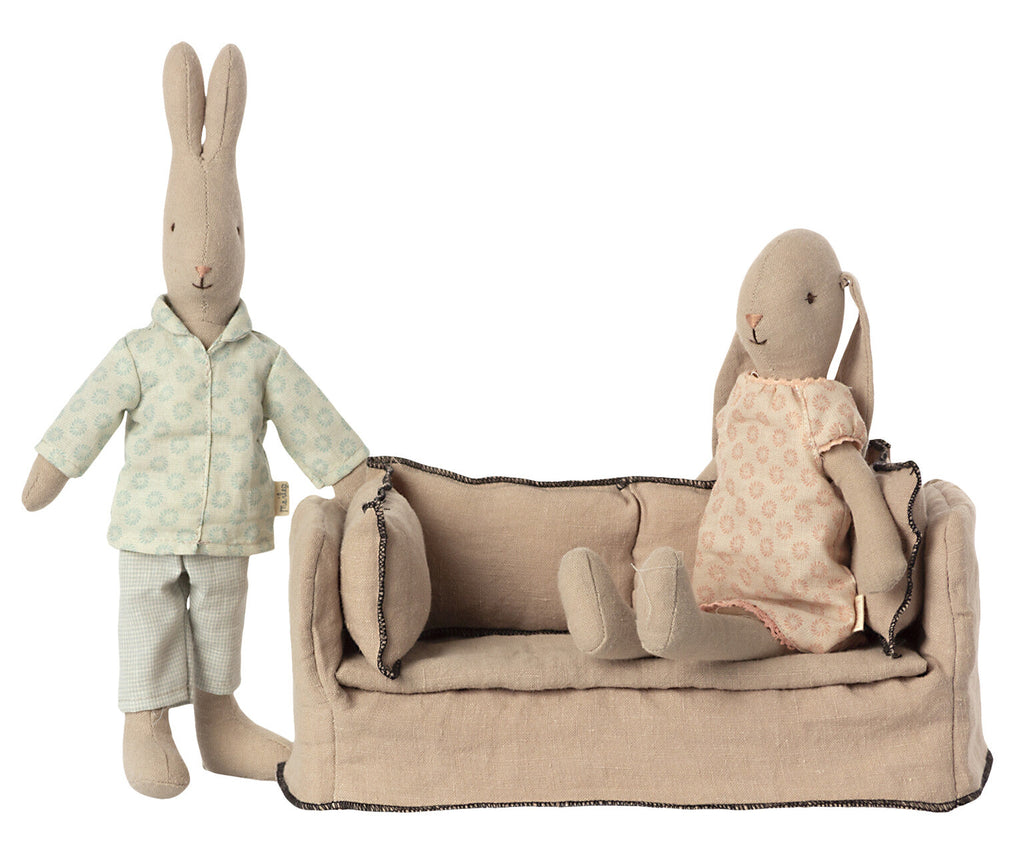 Two plush rabbit toys, one standing in a green patterned shirt and pants, the other sitting on a Maileg Dollhouse Couch, wearing a pink patterned dress.