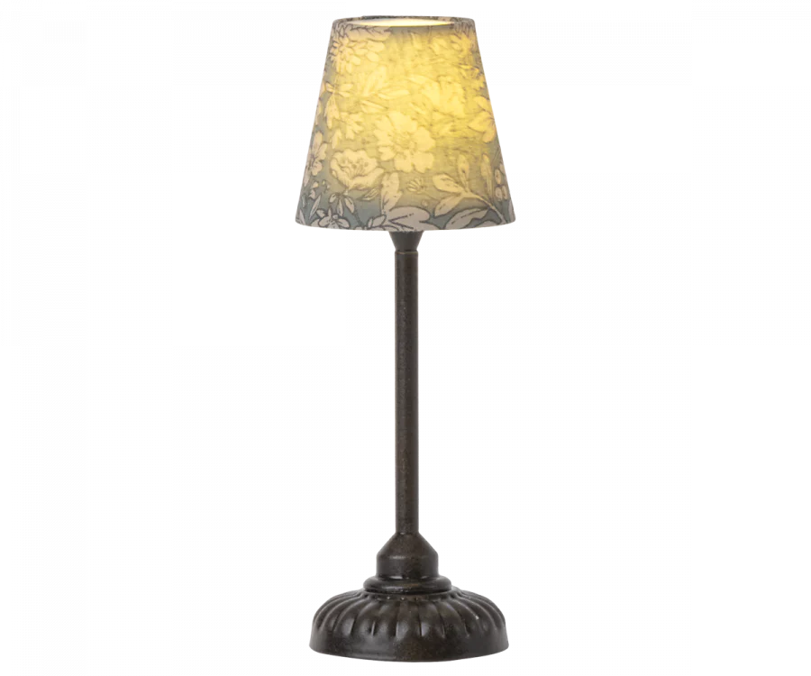 A table lamp with a dark, ornate metal base and a cone-shaped, yellowish shade decorated with floral patterns. The Maileg Vintage Mouse Lamp - Small is illuminated, casting a warm glow.