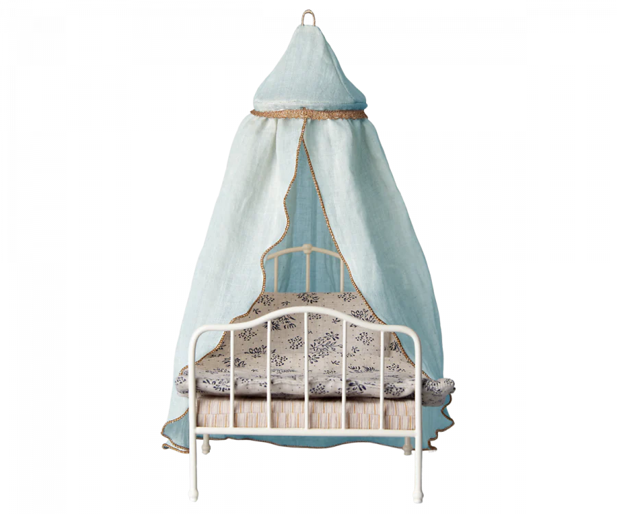 A small white metal-framed bed with a blue canopy draped over it creates an inviting retreat. The bed decoration features patterned bedding with white and blue designs. The Maileg Mini Bed Canopy flows down from the bedroom ceiling, providing a cozy, tent-like space for relaxation.