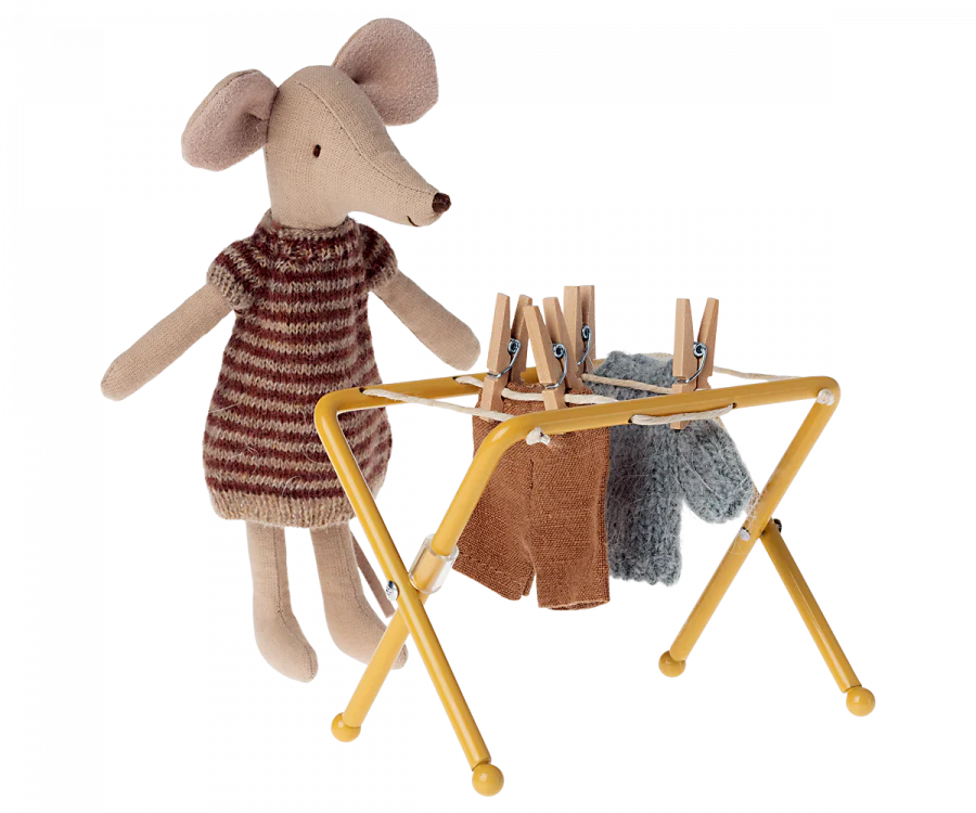 A plush mouse from the mouse family is dressed in a knitted dress, standing beside a small yellow Maileg Drying Rack, Mouse Size. The rack holds two pieces of doll-sized clothing secured with small wooden pegs.