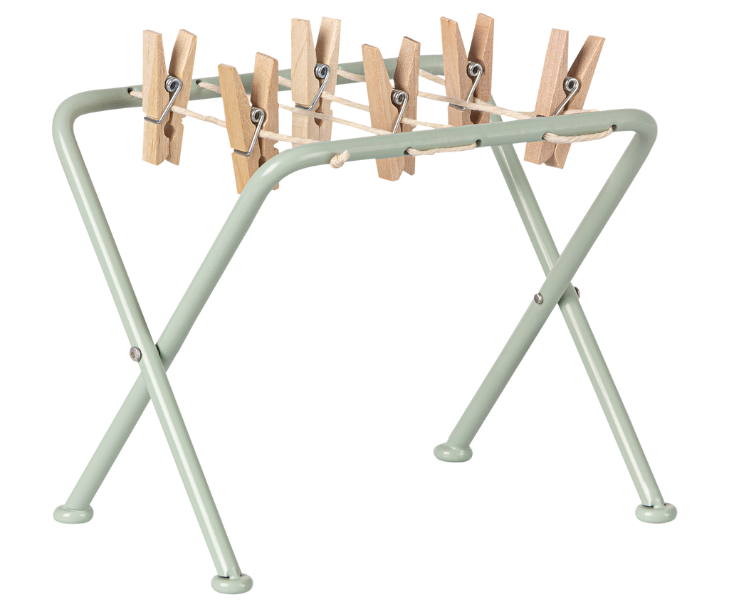 A small, light green metal Maileg Miniature Drying Rack featuring four wooden clothespins attached to a string stretched across its top. The Maileg Miniature Drying Rack has a simple, foldable design with crossed legs for stability, making it perfect for dollhouses or organizing small parts.
