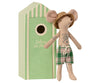 A Maileg Beach Mouse - Dad doll dressed like a tourist, with a straw hat and plaid swim trunks, standing next to a green, striped beach house labeled "cabine de plage.