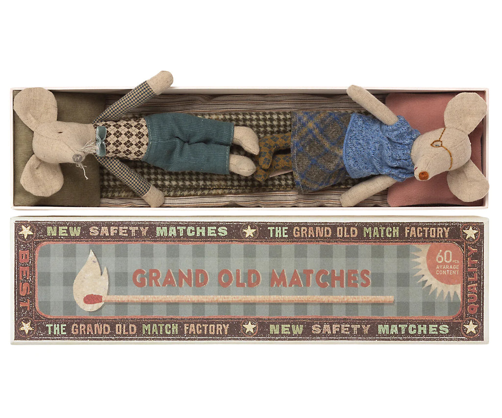 A whimsical display featuring two Maileg | Grandma & Grandpa Mice in Matchbox, one dressed in green and the other in blue, lying in a matchbox labeled "grand old matches" from "the grand old match factory.