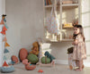 A toddler girl in a floral dress stands in a playroom filled with toys, including cuddly Maileg | Small Gantosaurus in egg - Green and colorful wooden toys, looking curiously towards a shelf.