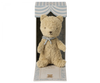 A beige teddy bear with a light blue scarf is displayed in a decorative box. The box, perfect as a new born baby gift, has a striped blue and white design with a window showcasing the bear. The text "Maileg My First Teddy, Sand" is visible on the box, making it a classic baby keepsake.