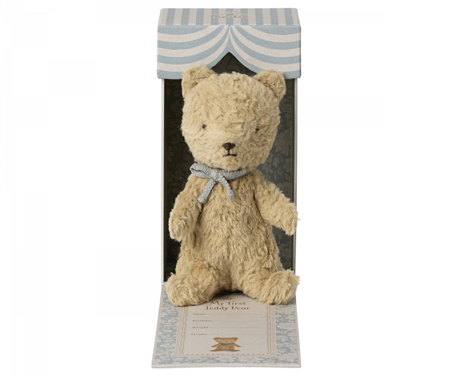 A beige teddy bear with a light blue scarf is displayed in a decorative box. The box, perfect as a new born baby gift, has a striped blue and white design with a window showcasing the bear. The text "Maileg My First Teddy, Sand" is visible on the box, making it a classic baby keepsake.