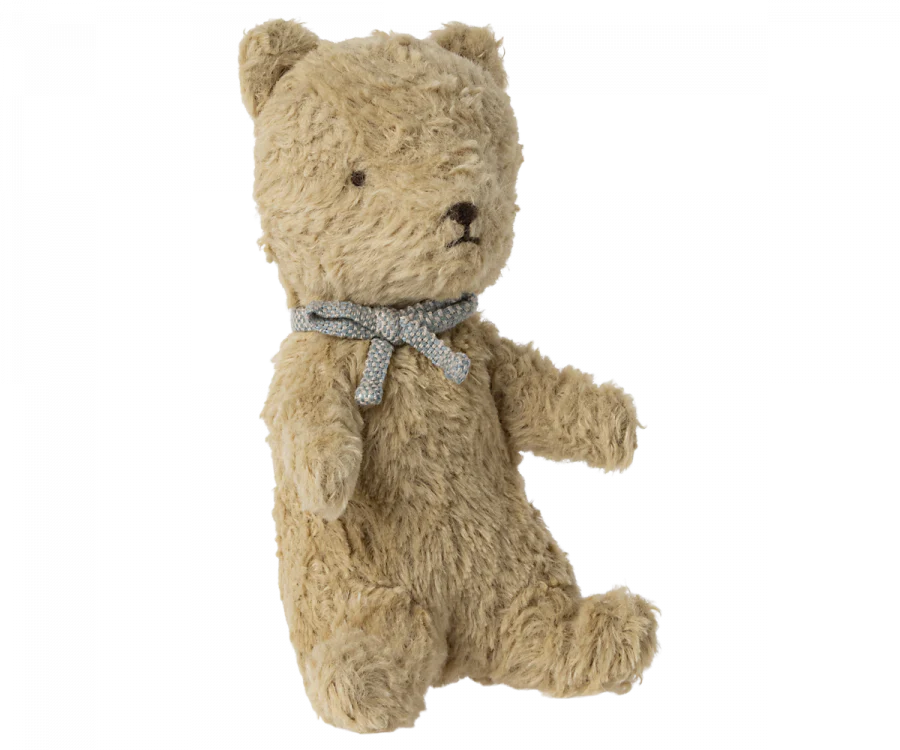 A small, beige Maileg My First Teddy, Sand with a fuzzy texture sits upright. It has round, dark eyes, a small dark nose, and a neutral expression. The teddy is wearing a light blue ribbon tied in a bow around its neck—a perfect classic baby keepsake for any new born baby gift.