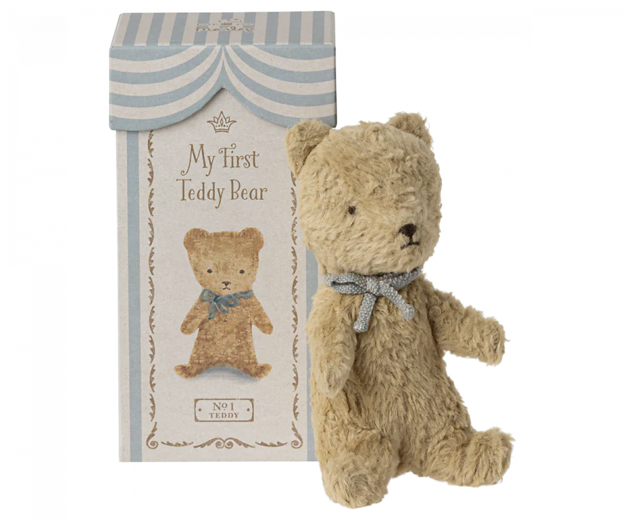 Image of a stuffed teddy bear next to a decorative box. The box's header reads "Maileg My First Teddy, Sand" and features an image of the bear adorned with a small gray ribbon around its neck. This classic baby keepsake comes in a striped blue and white top, perfect as a new born baby gift.