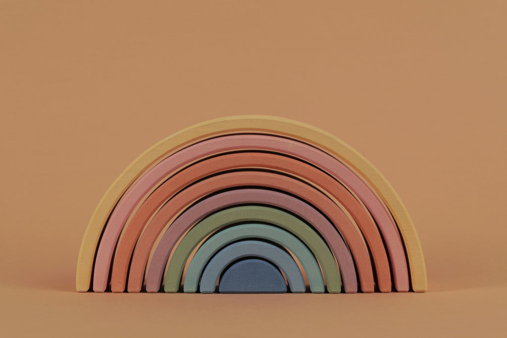 A Raduga Grez 9-Piece Rainbow Stacker wooden toy with graduated arches painted in a palette of pastel colors using non-toxic water-based paint, arranged against a soft peach background.