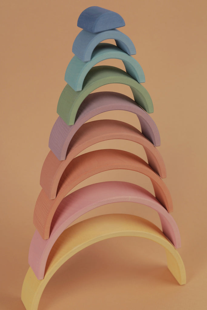 A stack of colorful Raduga Grez 9-Piece Rainbow Stacker arches in pastel shades, painted with non-toxic water-based paint, arranged in ascending order against a soft peach background.