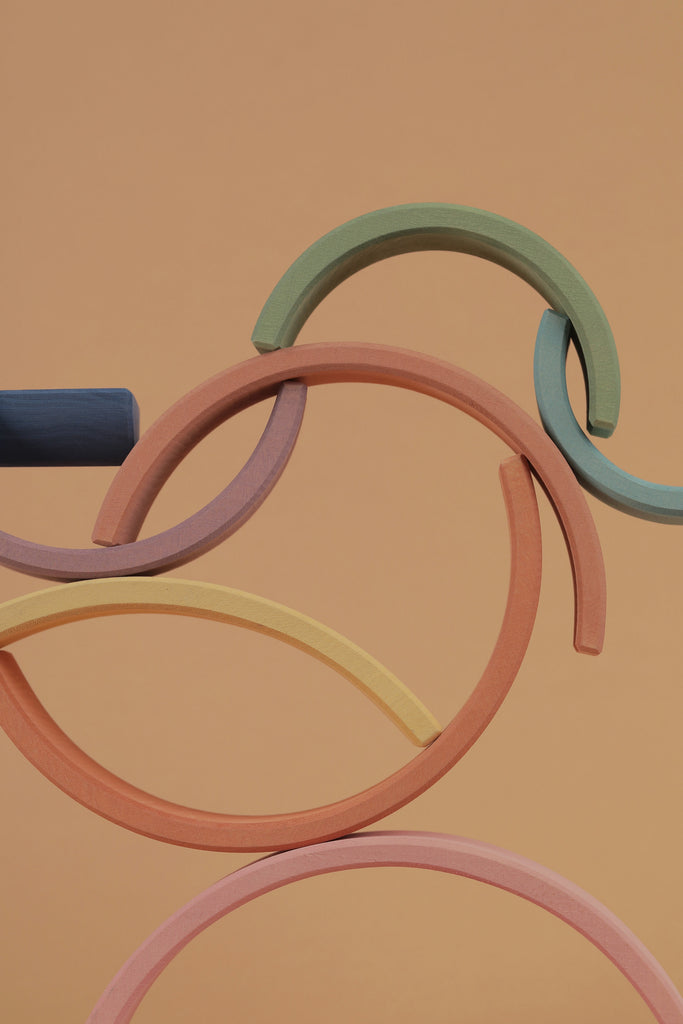 Colorful wooden curves, painted with non-toxic water-based paint, interlock gracefully against a peach background in an abstract and artistic arrangement with the Raduga Grez 9-Piece Rainbow Stacker.