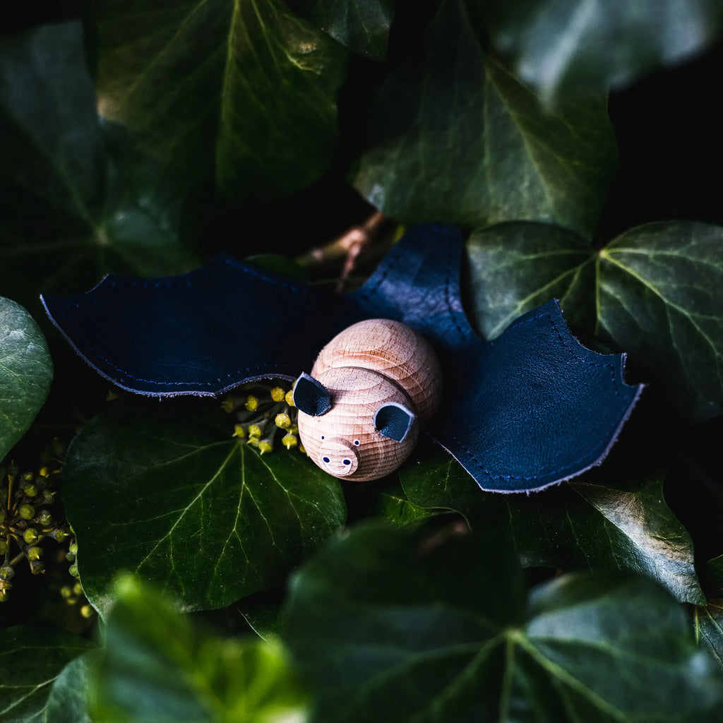 A charming hand-painted beech wood pig toy nestled among vibrant green ivy leaves, with tiny yellow blossoms scattered around and a Dark Blue Handmade Wooden Bat with leather wings in the background.
