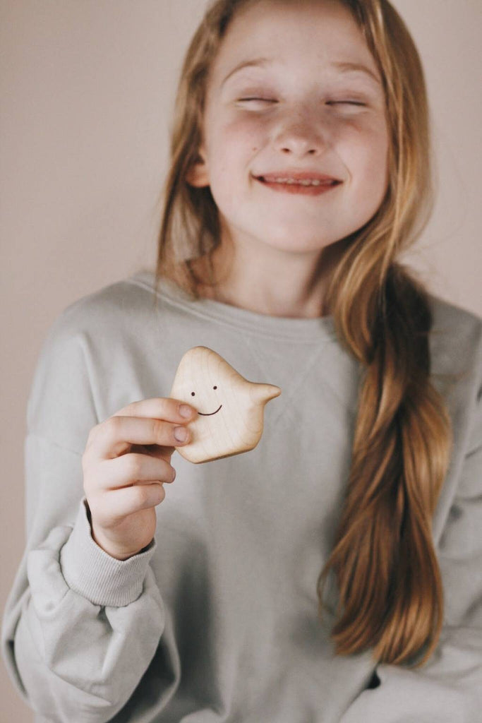 A girl with long, light brown hair is smiling with her eyes closed. She is wearing a light gray sweatshirt and holding a small Wooden Ghost Emotions toy crafted from natural linseed oil. The scene captures pure basic emotions of joy and serenity.