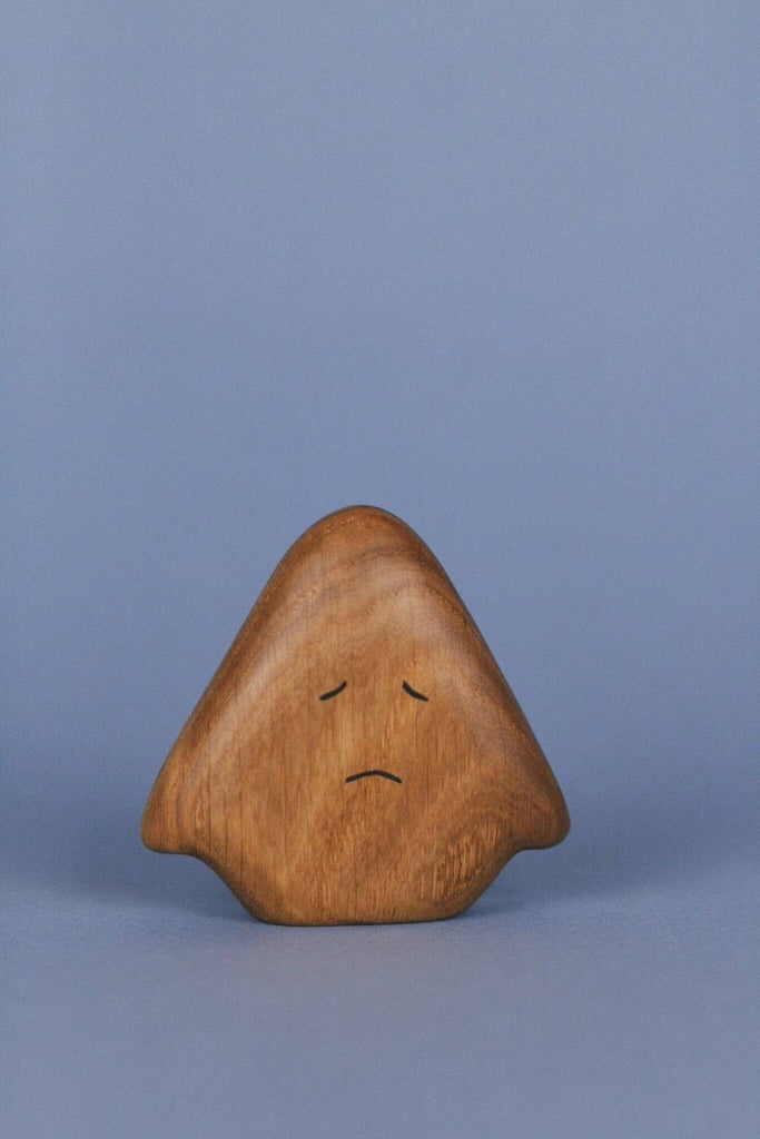 A small, smooth wooden figure with a rounded, triangular shape is set against a solid blue background. The Wooden Ghost Emotions has simple, etched facial features showing a sad expression with downward slanting lines for eyes and a frowning mouth.