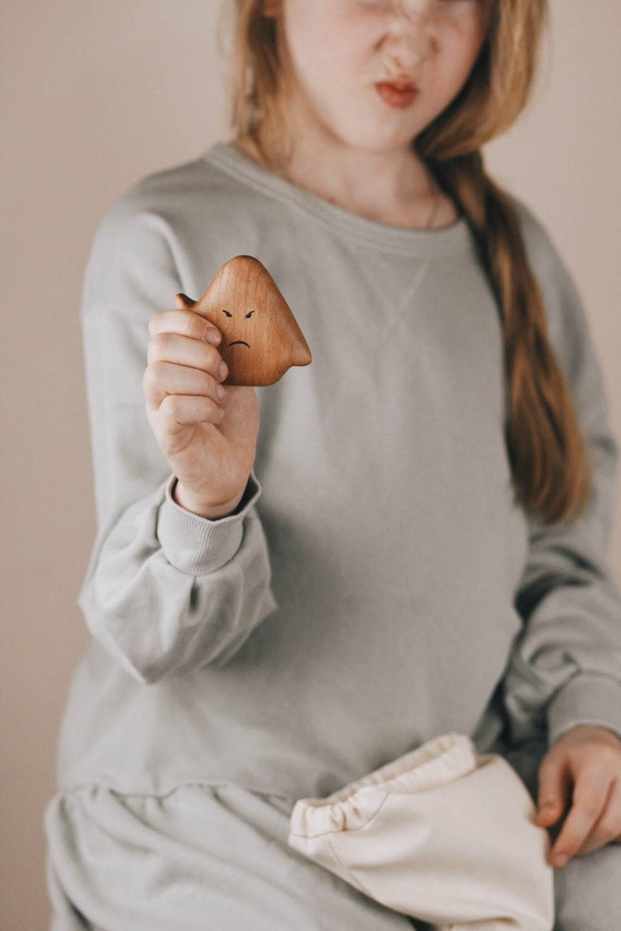 A child with long hair, wearing a gray long-sleeve shirt, holds a small wooden toy shaped like a rounded triangle with a sad face drawn on it. The toy, likely one of the Wooden Ghost Emotions, is coated in natural linseed oil. The child has a displeased expression and is also holding a light-colored cloth bag.