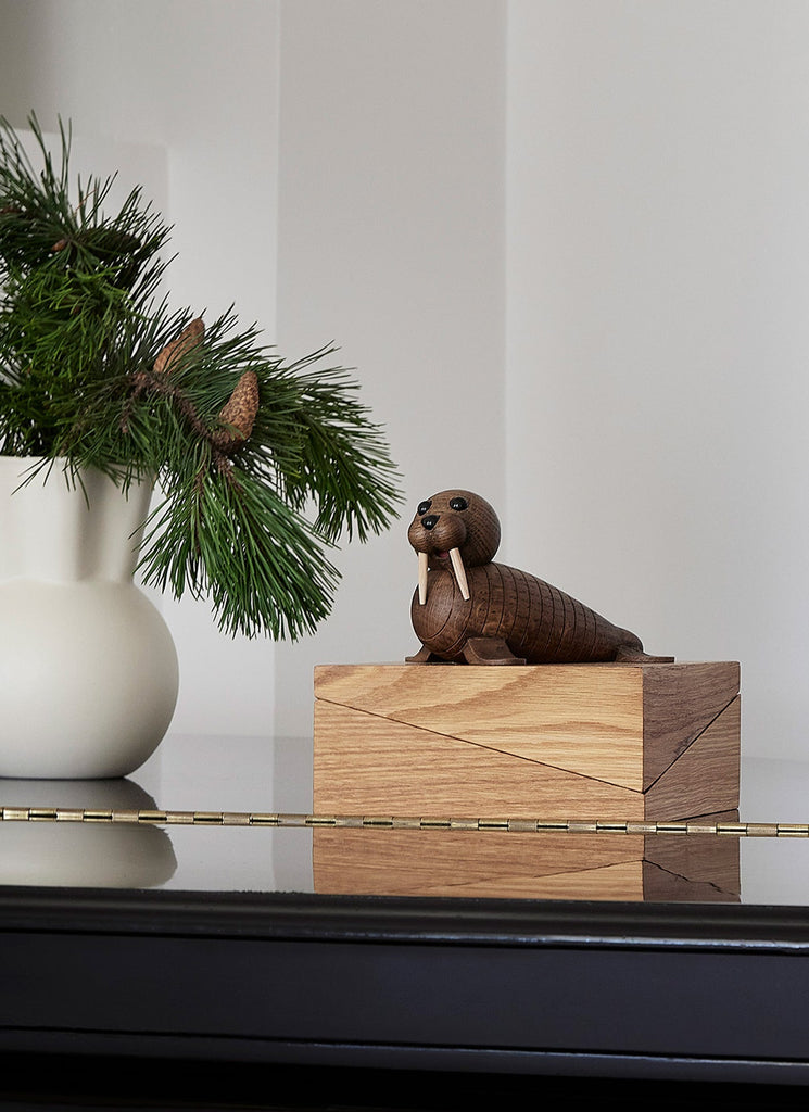 A handmade wooden Spring Copenhagen Wally figurine sits on a geometric block on a piano, under a branch of pine with pine cones in a minimalist vase, suggesting a serene, styled interior setting.